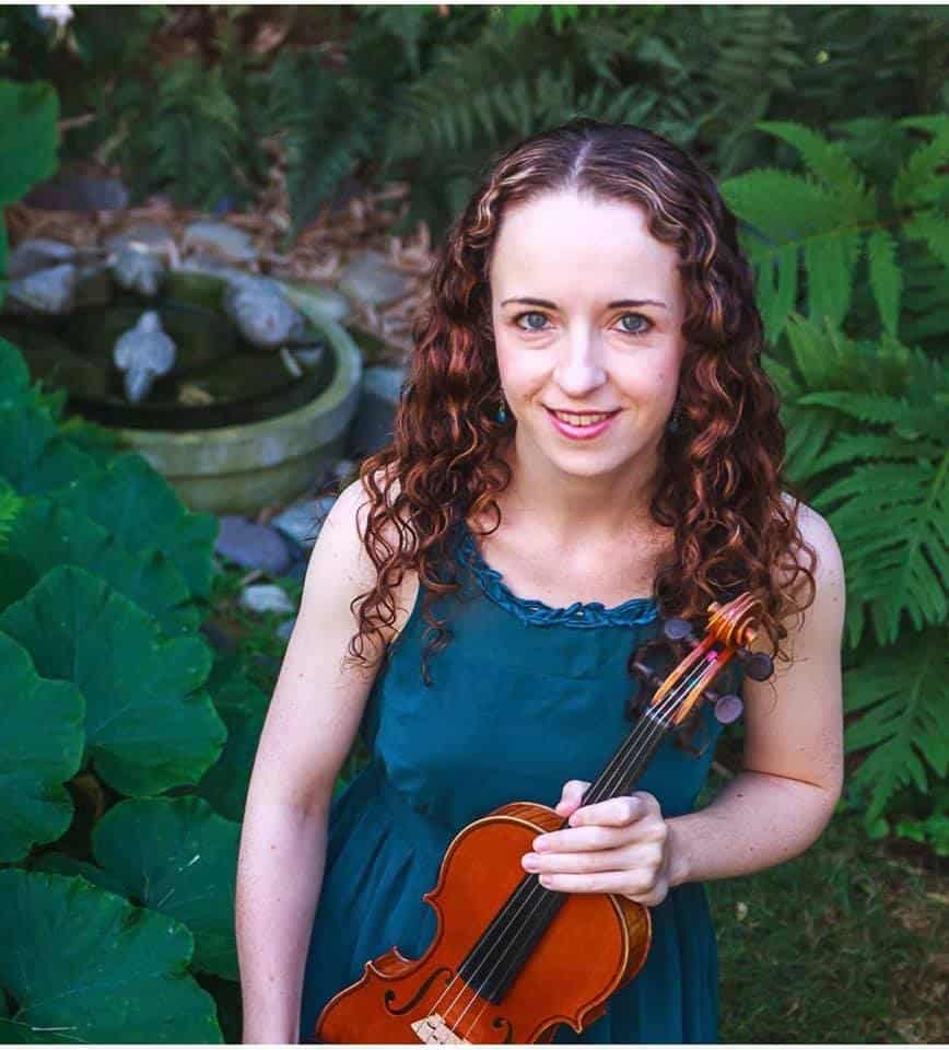 Violinist stands in the foreground behind a verdant backdrop of plants and a fountain
