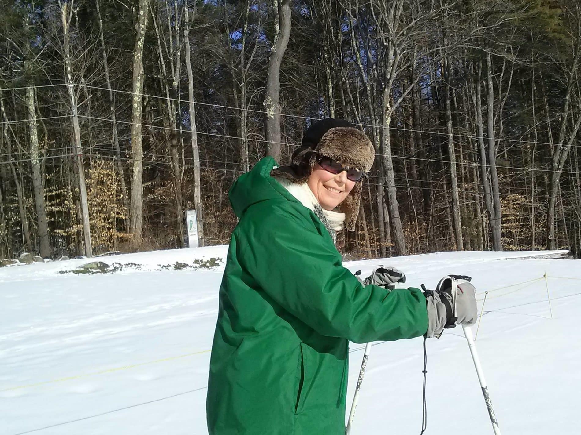 A cross country skier smiles at the camera in her green jacket while she makes her way through the snow