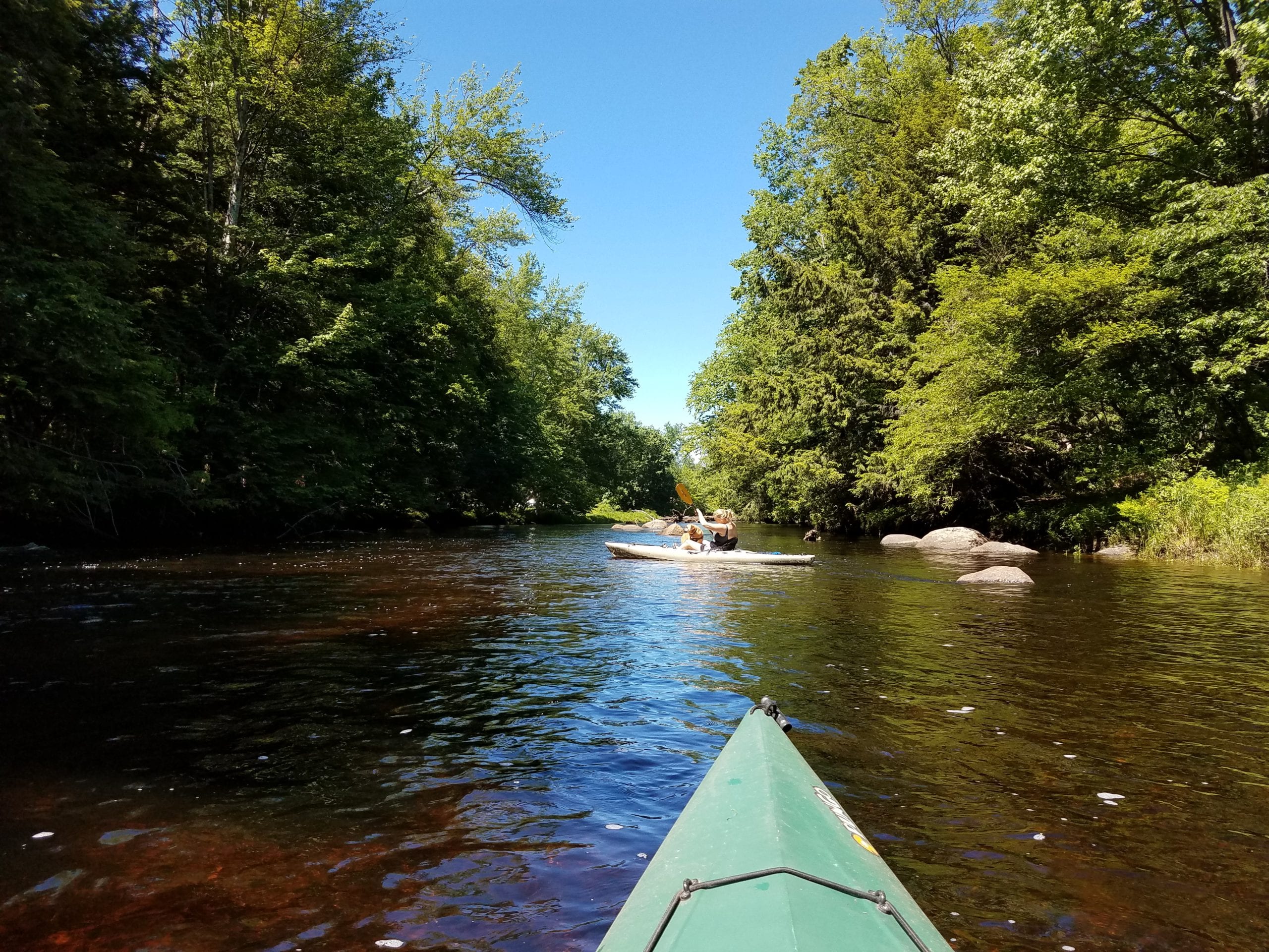 The tip of a kayak is visible at the bottom of the image on a river. Ahead is another person kayaking with green trees on each side.