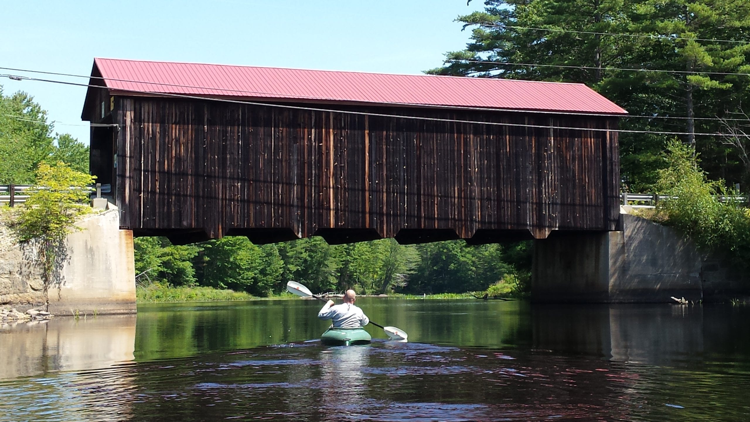A covered bridge sits in the background while a kayaker paddles toward it to go underneath