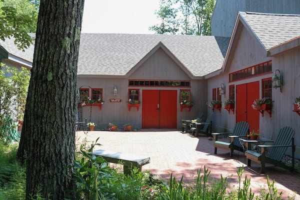 An old farmhouse converted into a theatre is painted gray, with red doors. A pathway leads from the foreground toward the threatre, while a tree trunk takes up the left half.