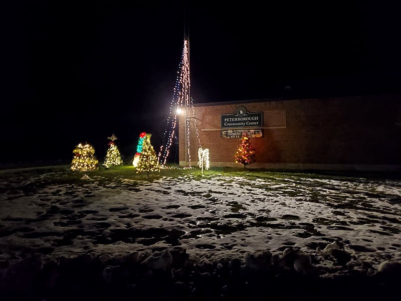 A brick building in the background with a sign that says 'Community Center'. In the foreground a pole with lights draped outward and several christmas trees are decorated.