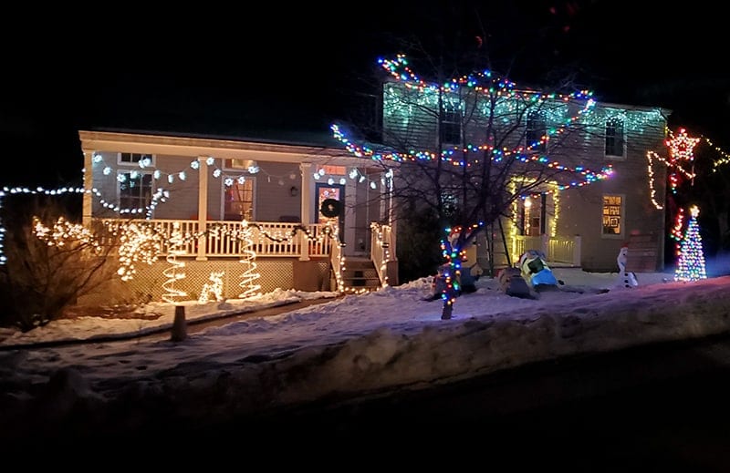 A house with lights made out of trees in the front of their deck. Snowflake lights hang from the trim, and a tree is decorated in multicolor lights
