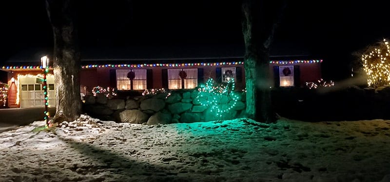Blue lights hang from a small tree in front of the house, while more hang from the roof. Each window has a small wreath on it
