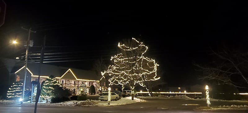 holiday lights strung up on trees and bushes in the foreground with the inn lit up with lights on the trim of the roof.