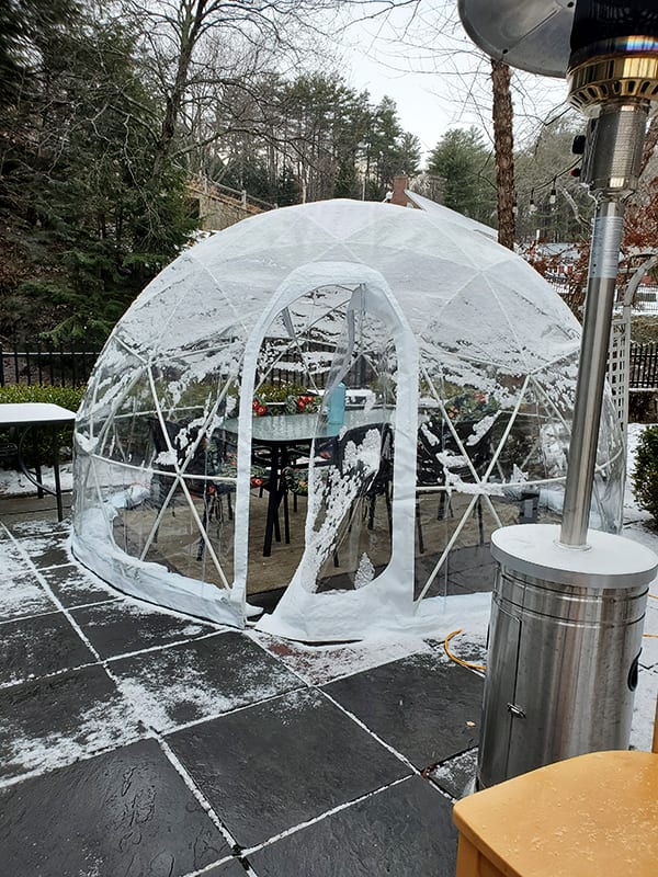 clear dome plastic igloo surrounds an outdoor dining table with snow nearby