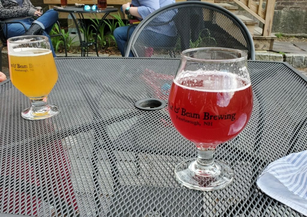Two beer glasses with stems sit on a wire table. One beer is a reddish hue while the one behind it is more golden.
