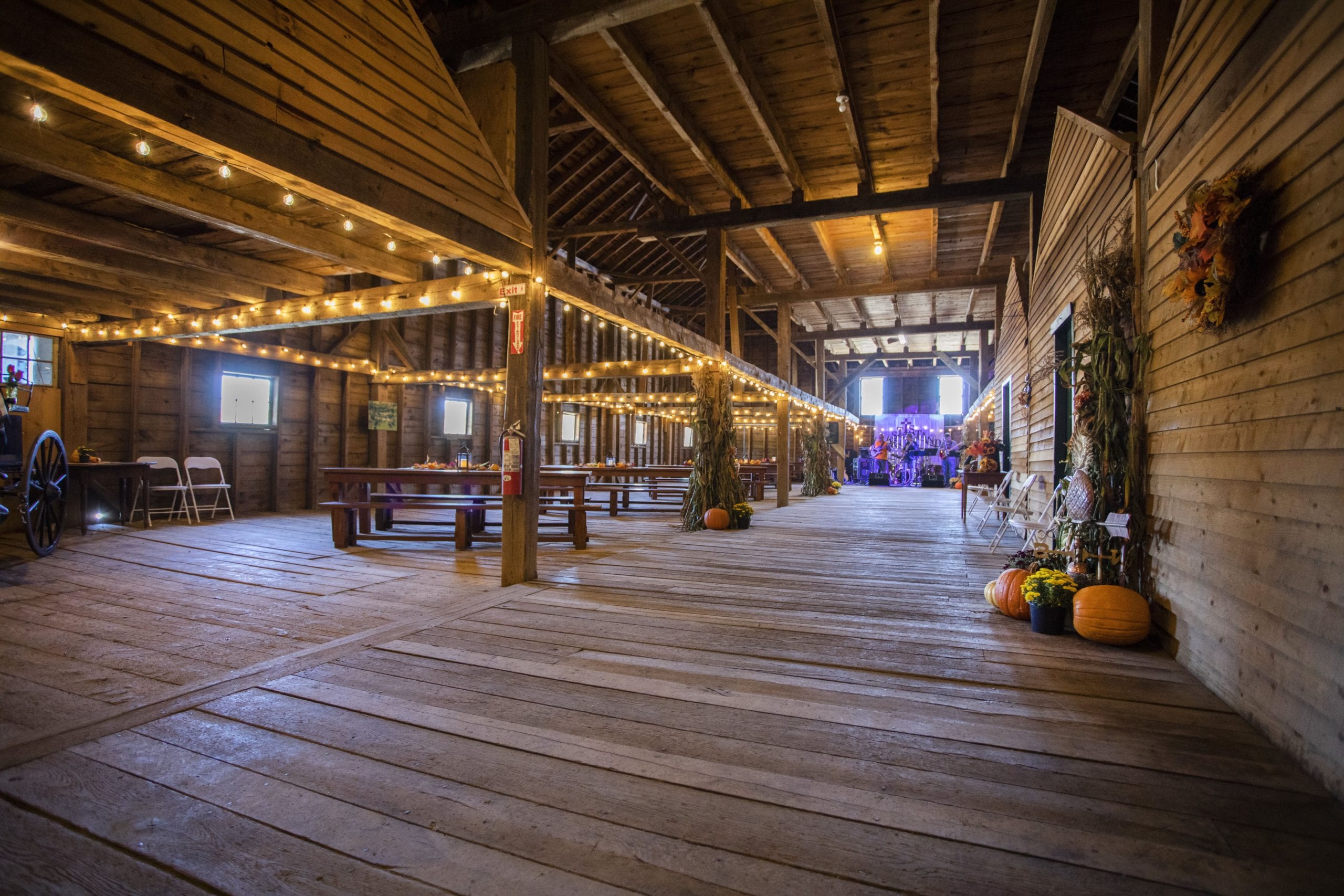 Interior of a barn that has been converted. Bulb twinkle lights hang above low beams on the left where tables are arranged, while the far end has large windows overlooking the inside.