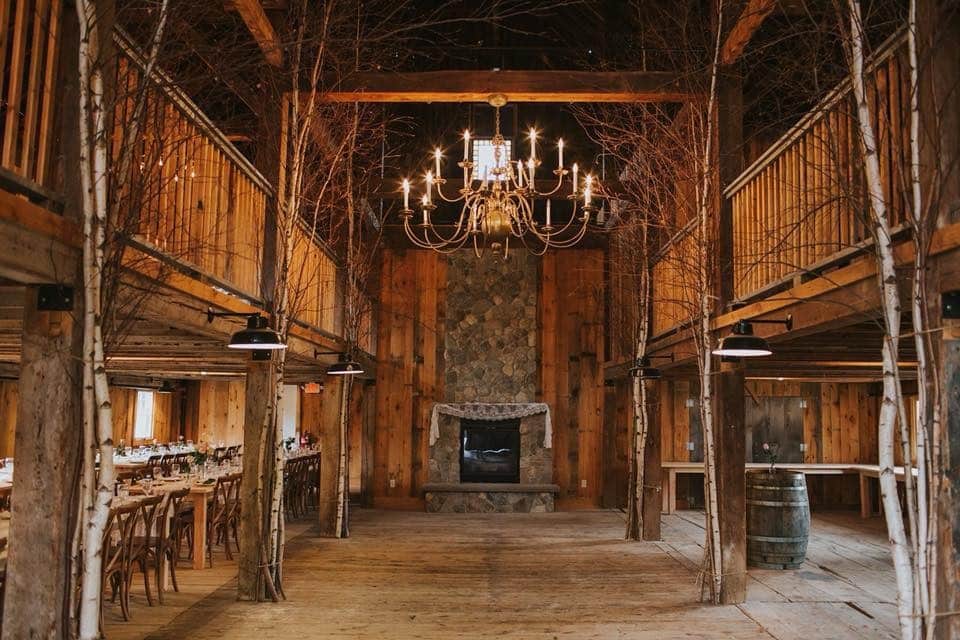 Interior of a wooden barn's middle aisle. Rows of bench seats and tables on either side, a chandelier above, and a stone fireplace in the center back.