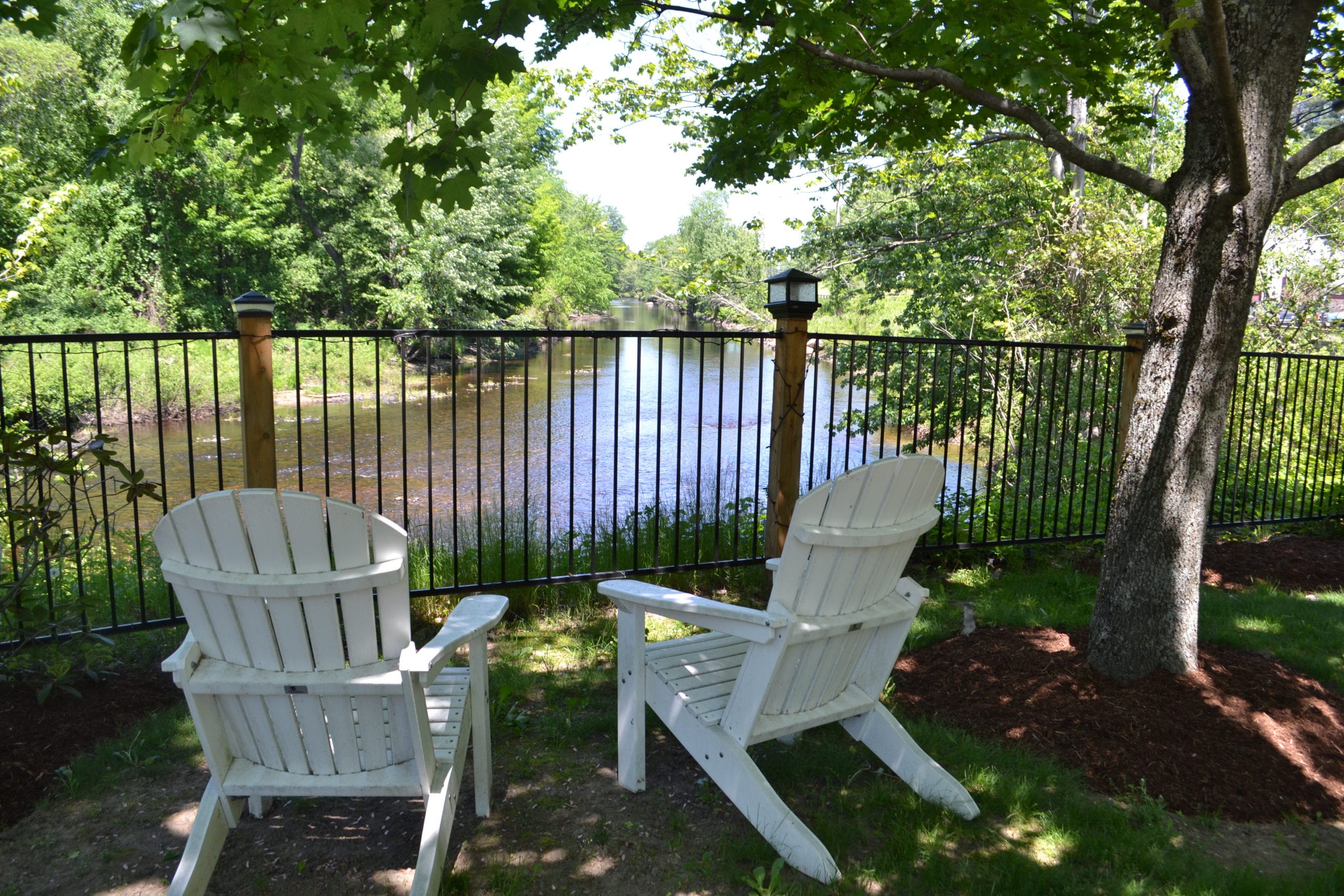 Two white adirondack chairs face the fence separating the river from the grassy area the chairs sit on