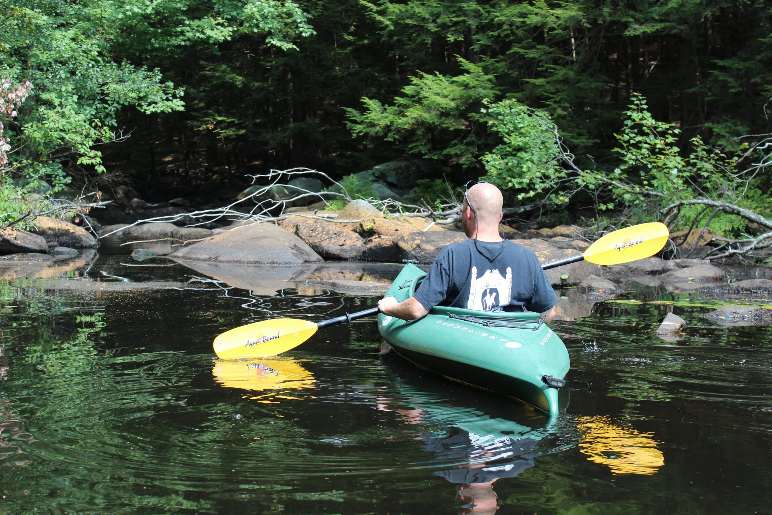 A kayaker maneuvers his kayak on the water. Rocks can be seen head on the edge of the waterway.
