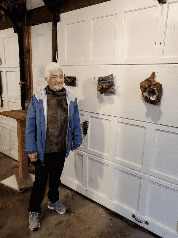 Artist Ann Falby stands to the left of her sculptures that hang on the wall to her right.