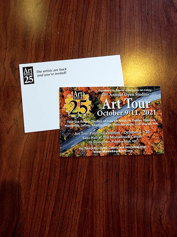 A postcard of a road through fall foliage with the 25th Annual Art Tour's information faces upward, with the white back of another postcard behind it