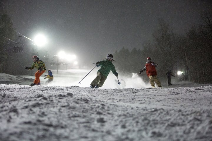 A dark ski slope with lights shining above highlights several skiers and snowboarders coming down the slope.