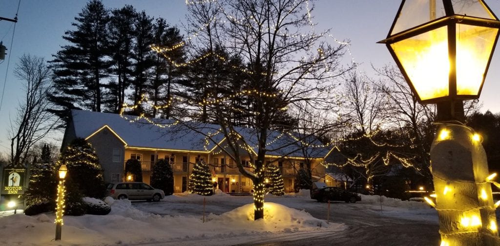 As sunsets in the background the Jack Daniels Inn can be seen with snow on the roof and ground. Lights line the hotel and the trees as well as the lamp post to the direct right of the view.