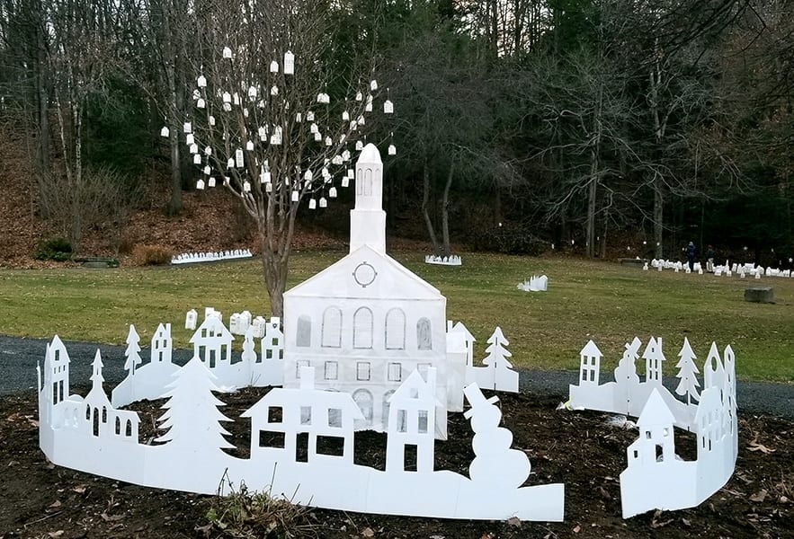 A church surrounded by trees and houses cut out of white paper arranged in a circle inside a park. In the background smaller houses that serve as lanterns are made out of the same paper.
