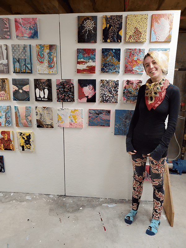 Artist Lauryn Welch stands in front of a studio wall with rows of small square artwork.