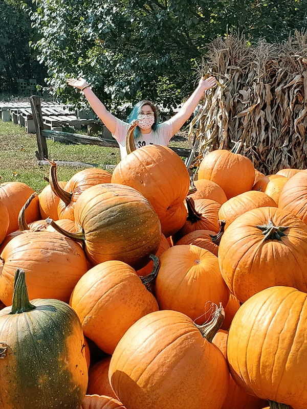 A pile of pumpkins in the foreground, with a woman that has her arms raised above her head stands behind them