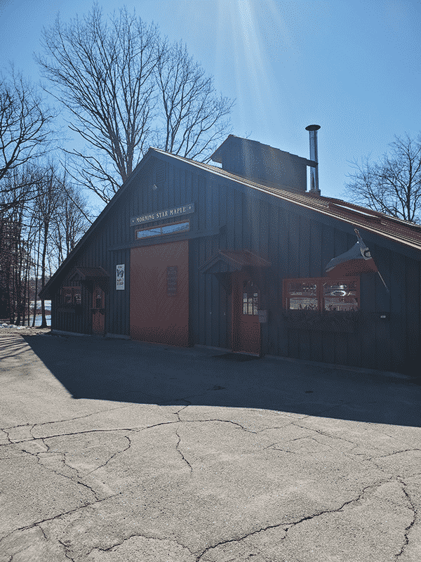 A dark gray building with a red roof sits in the forefront with clear skies in the backdrop. A sign that reads Morning Star Maple hangs on the front