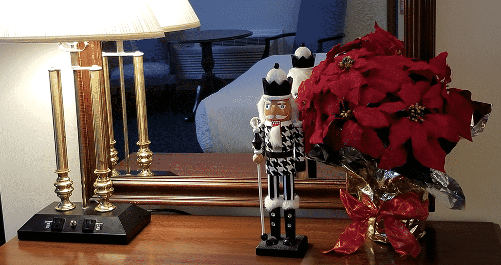 A desk in front of a wall mirror with a lamp to the left and a black and white checkered nutcracker in the center. To the right of the nutcracker is a vibrant poinsettia.