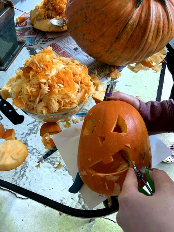 A pumpkin is being held by the carver as they use the potato peeler to round out the edges of the mouth. On the table a pile of pumpkin guts sits.