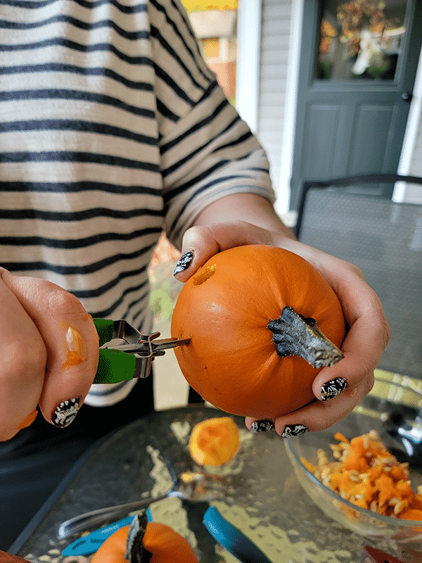 Person carving uses a potato peeler to make round eyes in a tiny pumpkin