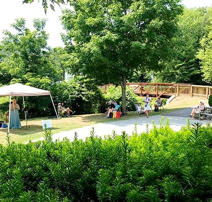 A shot of a pathway from a foot bridge to the right shows musicians performing under a tent while listeners sit on a bench nearby